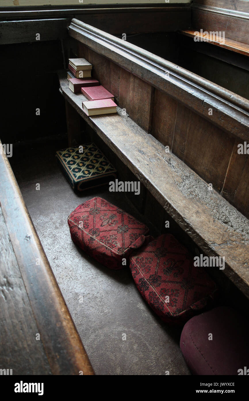 Alte Kirche pew durch Fenster Licht beleuchtet läuft diagonal über dem Rahmen: gestapelte gesangbücher an einem Ende und rot, grün gemusterten Kniebänke unter. Stockfoto