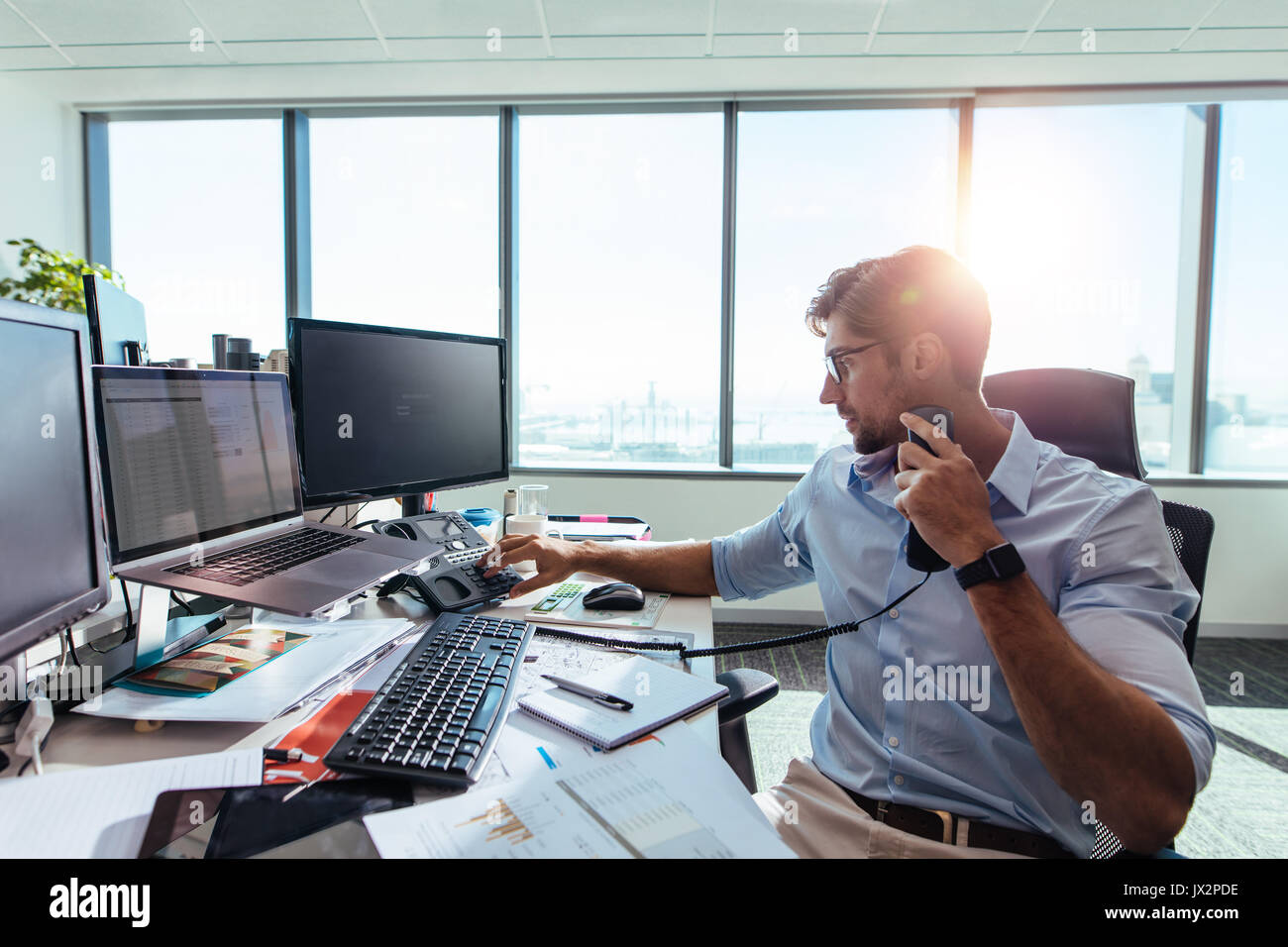 Geschäftsmann, der in seinem Büro arbeitet, mit Computern und Geschäftspapieren auf dem Tisch. Junger Unternehmer, der einen Anruf über ein Telefon an seinem Bürotisch tätigt. Stockfoto