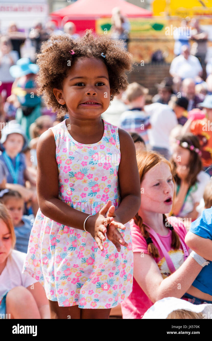 Afro-karibische Kind, Mädchen, 6-7 Jahre alt, die stehen im Publikum sitzen Kinder, trägt ein rosa Kleid mit Blüten. Intensive Mimik der Konzentration, als Sie versucht, zu klatschen. Stockfoto