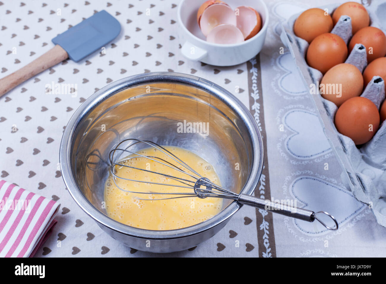 Geschlagenen Eier und eine Hand in einem Schneebesen aus Edelstahl Schüssel auf ein Land kitcken Tabelle und eine Schachtel Eier Stockfoto