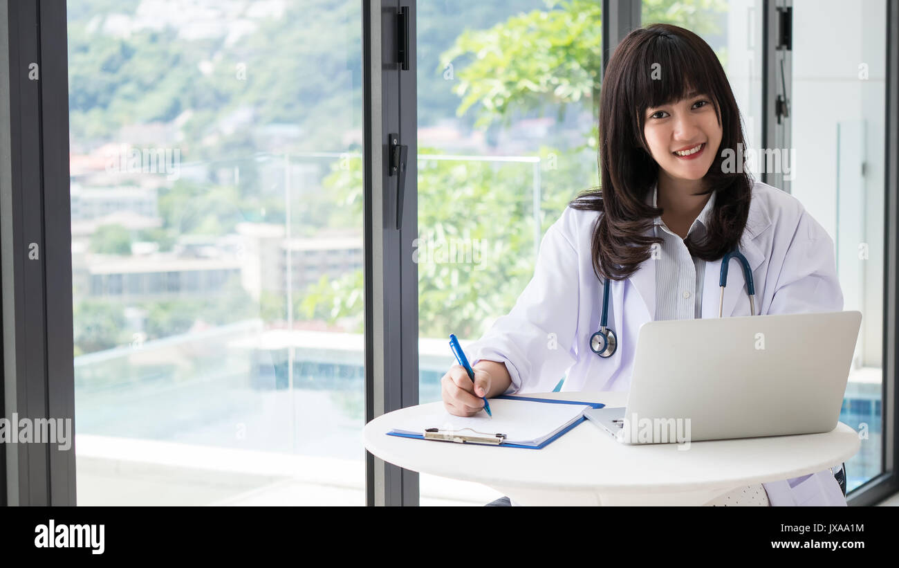 Junge Asien Arzt Lächeln und Schreiben einen Bericht für einen Bestrahlungsplan mit Stethoskop, die Hand am Stift und Informationen mit Laptops finden Sie in Stockfoto