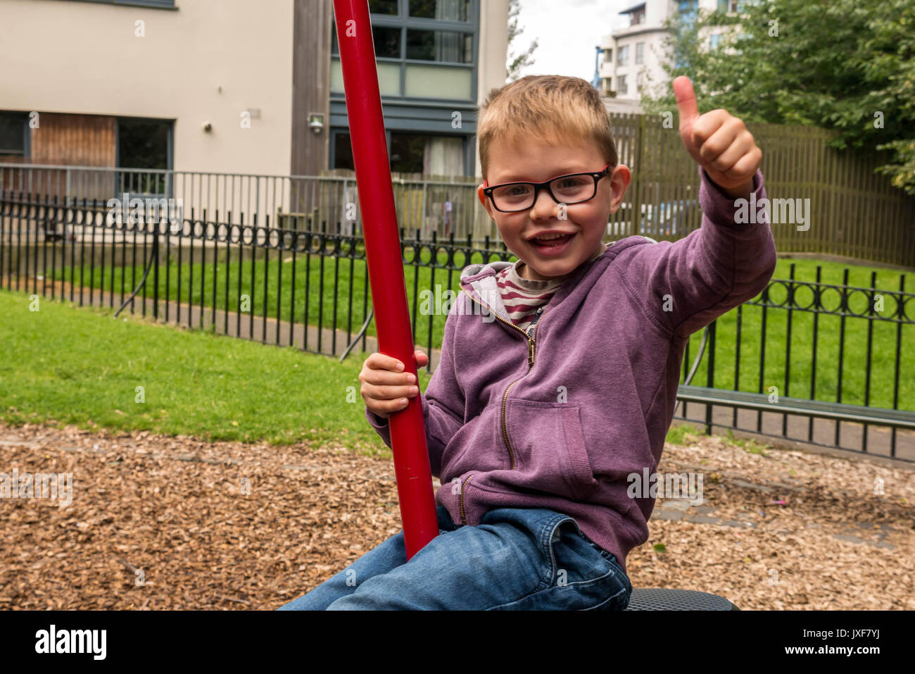 Nahaufnahme von Glücklich lächelnde Junge mit Brille auf einer Schaukel auf dem Spielplatz, die Daumen oben unterzeichnen, Leith, Edinburgh, Schottland, Großbritannien. Stockfoto