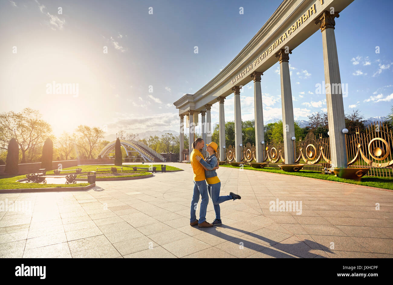 Glückliches Paar in blauen und gelben Tüchern umarmen in der Nähe von Spalte und Brücke im berühmten Park in Almaty, Kasachstan Stockfoto