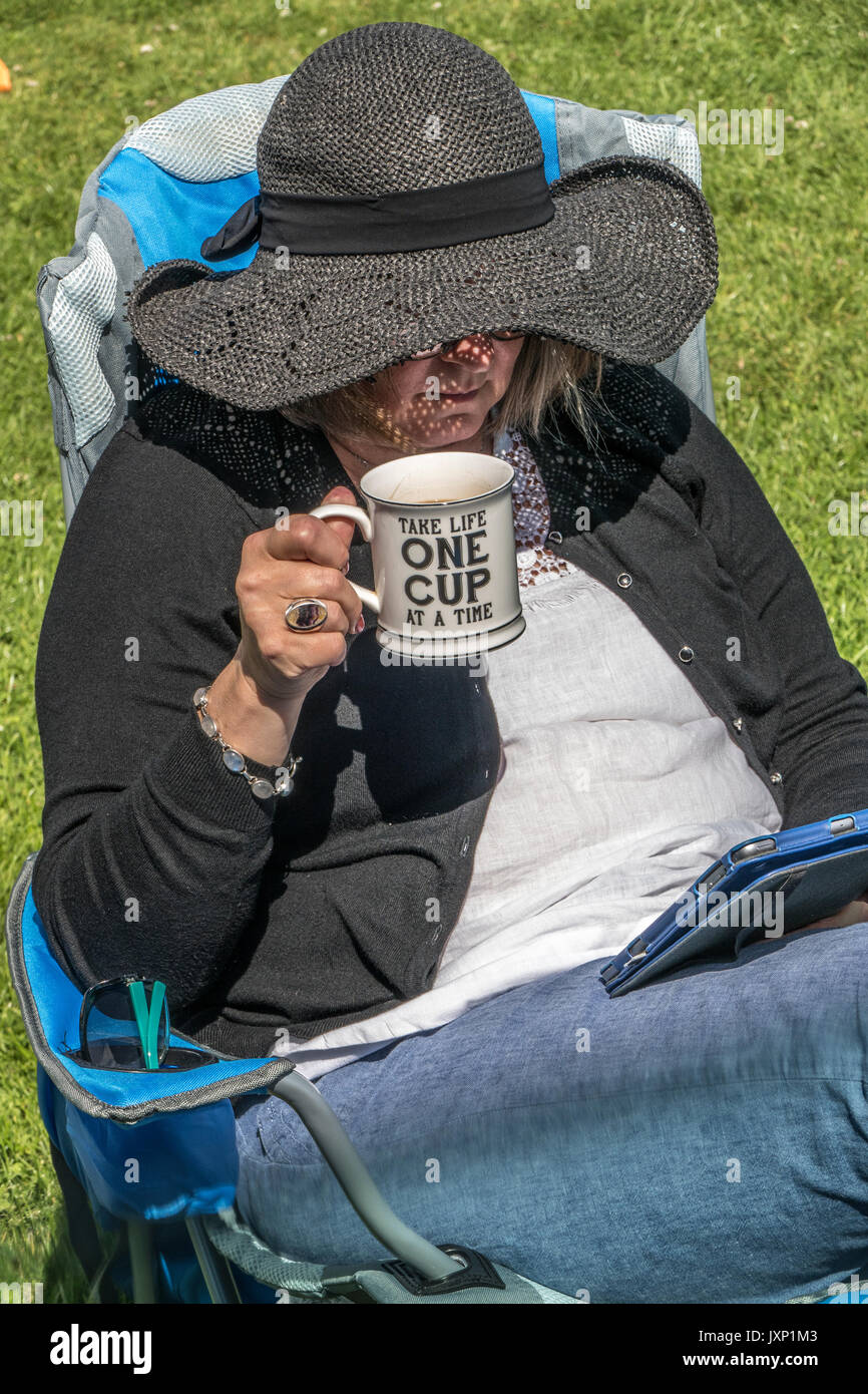 Eine Frau mittleren Alters trinken aus einem Becher, beim Sitzen in einer Camping Stuhl und über einen Tablet-PC. Stockfoto
