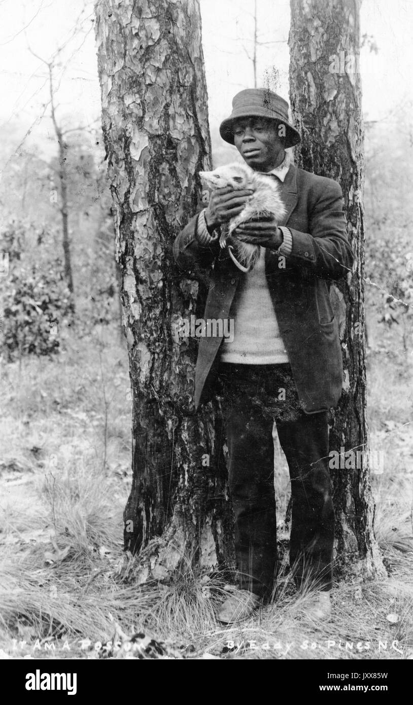 Ein reifer Afroamerikaner, der einen Hut und Mantel trägt, steht in einem bewaldeten Gebiet und schaut auf ein totes Possum, das er in seinen Händen hält, möglicherweise in South Carolina, 1918. Stockfoto