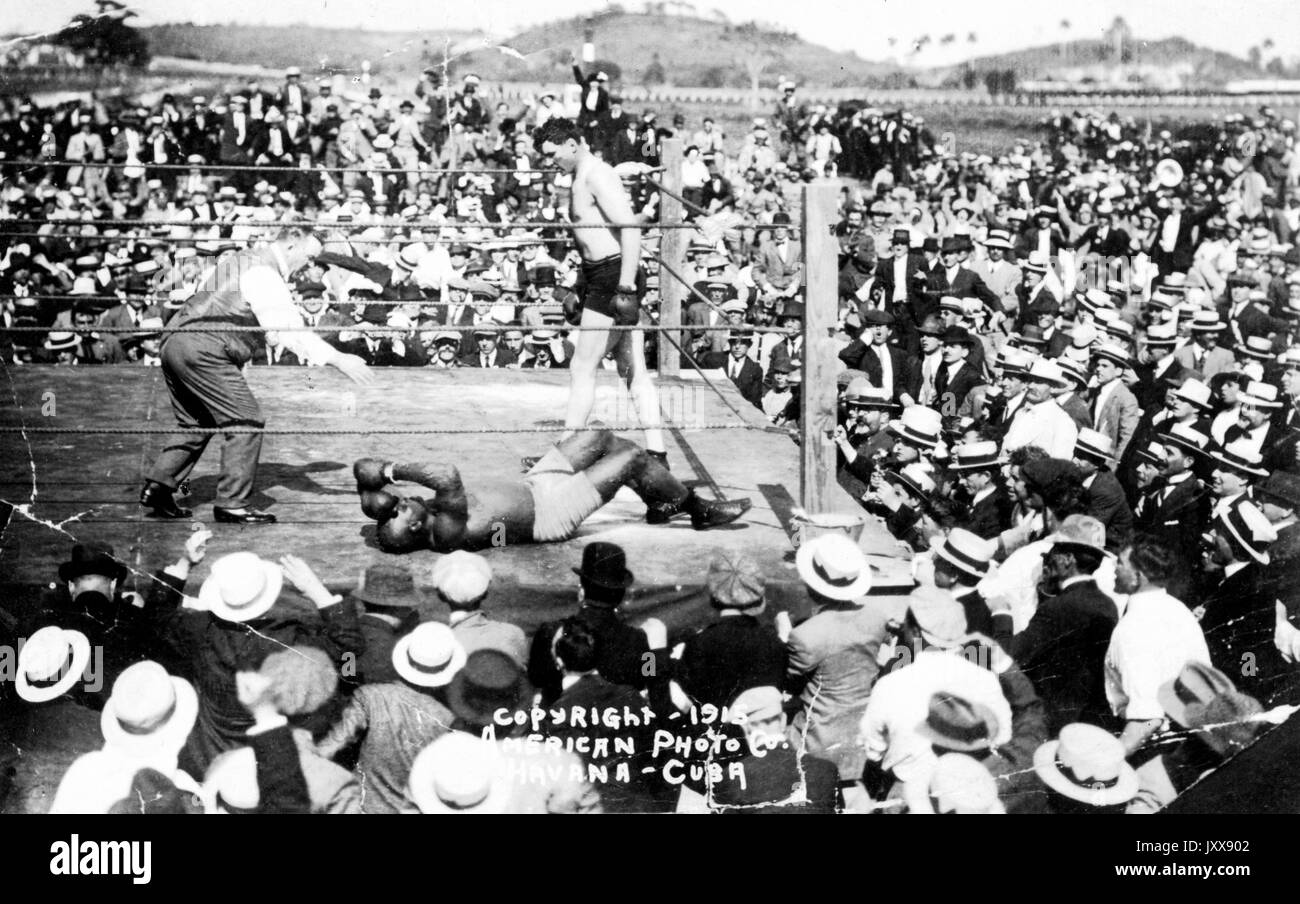 Eine große Gruppe von Zuschauern steht um einen Boxring im Freien, in dem Jack Johnson (rechts), ein Schiedsrichter (links) und Jess Willard (Boden), der gerade von Johnson in der 26. Runde geschlagen wurde, um die Schwergewichtsboxmeisterschaft in Havanna, Kuba, 1915 zu gewinnen. Stockfoto