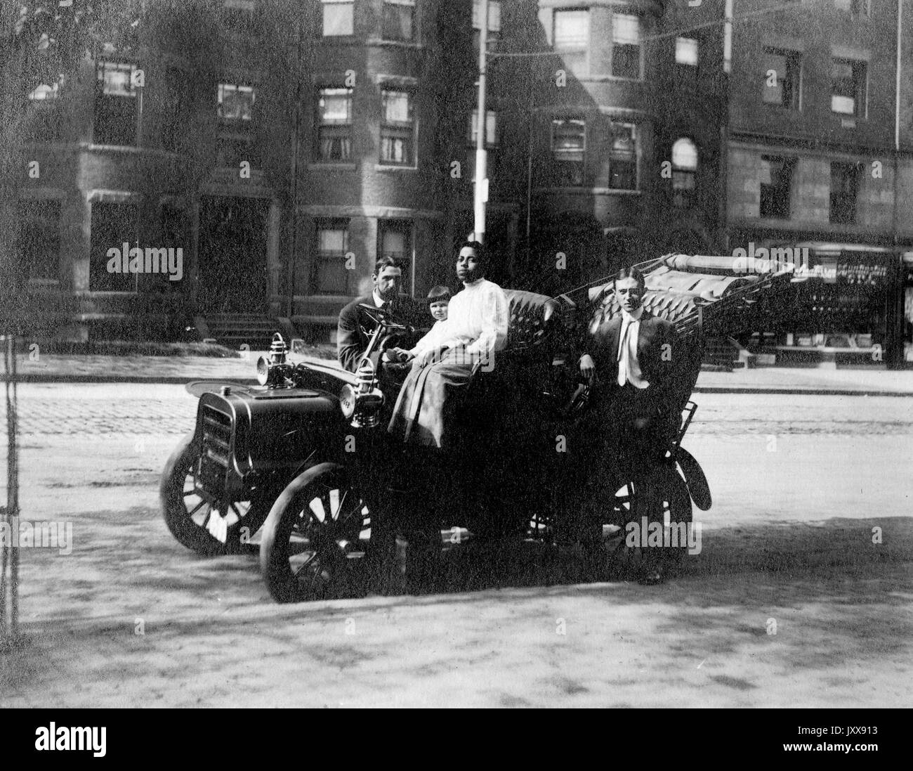 Ein reifer Kaukasischer Mann und junges Mädchen sitzen in einem Auto, während ein junger Kaukasischer Mann steht gegen die Seite des Autos und eine junge afroamerikanische Frau sitzt in der Fahrersitz des Autos, das in der Mitte einer Stadtstraße, möglicherweise Boston, Massachusetts, 1915 ist. Stockfoto