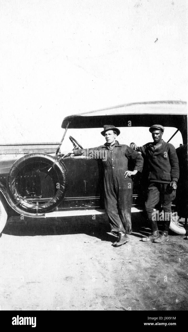 Ganzkörperporträt von zwei Männern, die bei einem Auto stehen, einem Kaukasischen, einem afroamerikanischen Mann, der Uniformen trägt, 1920. Stockfoto