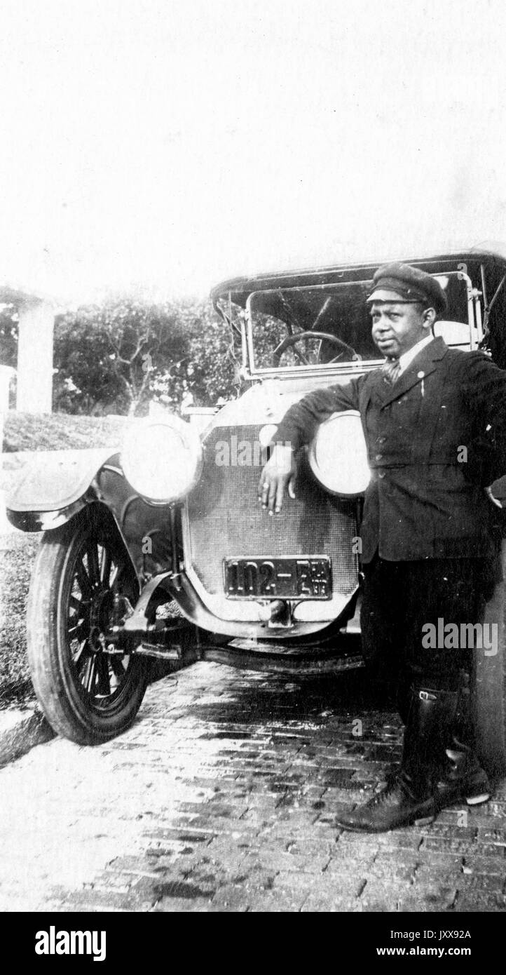 Ganzkörperporträt eines afroamerikanischen Mannes, der sich gegen die Scheinwerfer eines Autos lehnt, in Chauffeurs Uniform, neutraler Gesichtsausdruck, 1919. Stockfoto