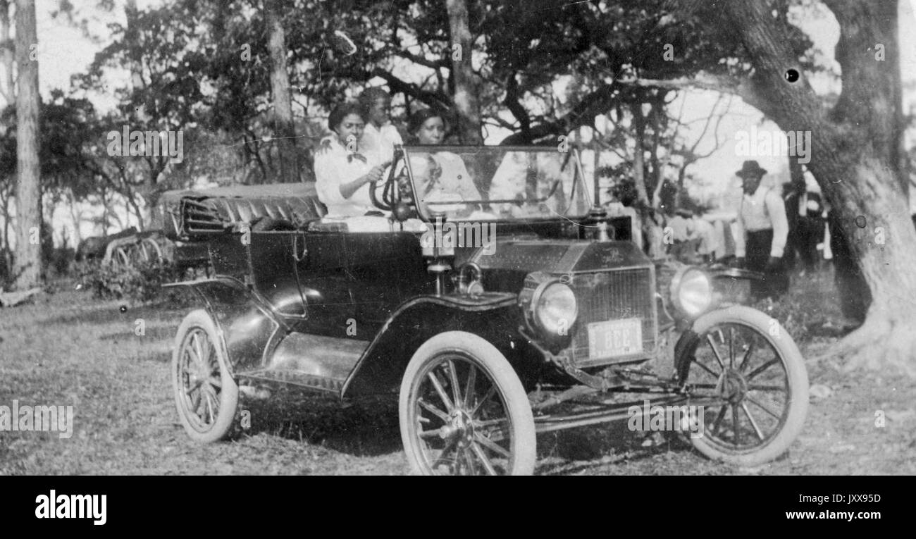 Porträt von drei afroamerikanischen Mädchen, die ein Auto im Freien auf Gras unter einem Baum fahren, ein Mann im Hintergrund beobachten, 1920. Stockfoto