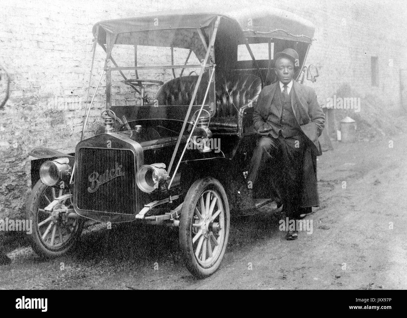 Ganzkörperaufnahme eines afroamerikanischen Mannes, der sich gegen ein Buick-Auto lehnt, das gegen ein Gebäude geparkt ist, einen Anzug und einen Hut trägt, der auf seinem Kopf geneigt ist, neutraler Gesichtsausdruck, 1920. Stockfoto