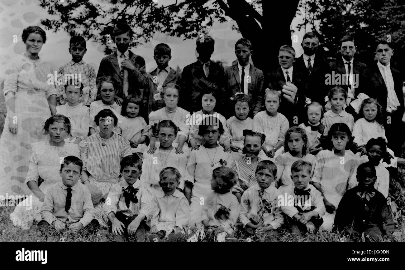 Landschaftsaufnahme von Schulkindern, alle im Gras unter einem Baum sitzend, eine Reihe stehend, alle Jungen und Mädchen in weißen Schuluniformen, ein afroamerikanischer Junge in der unteren rechten Reihe mit schwarzem Hemd, neutrale Gesichtsausdrücke, 1920. Stockfoto