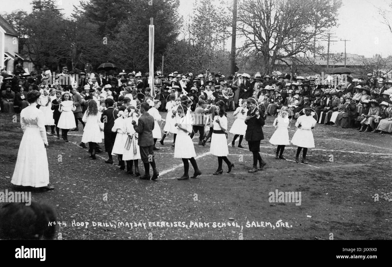 Lange Landschaftsaufnahme von Schülern, die im Rahmen der Mayday-Übungen einen Reihenkurz machen, Mädchen in weißen Kleidern, Männer in Anzügen, Erwachsene Zuschauer im Hintergrund; Salem, Oregon, 1920. Stockfoto