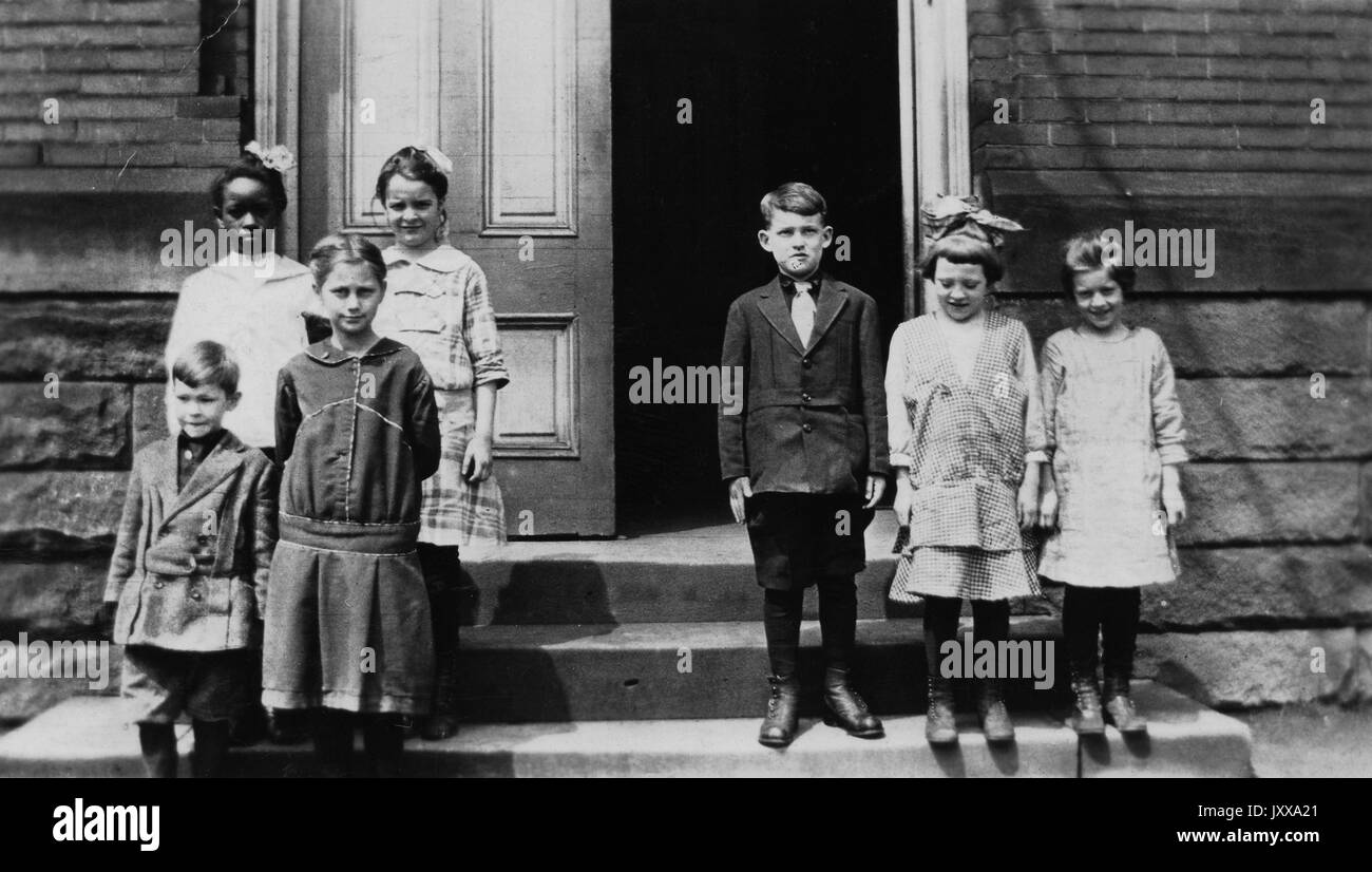 Ganzkörperportrait von zwei Kindergruppen; drei kaukasische Kinder, zwei Mädchen und ein Junge, auf der rechten Seite; vier Kinder, eines davon ein afroamerikanisches Mädchen, auf der linken Seite; großer Raum zwischen zwei Kindergruppen, stehend auf einer Treppe vor dem Gebäude, 1920. Stockfoto