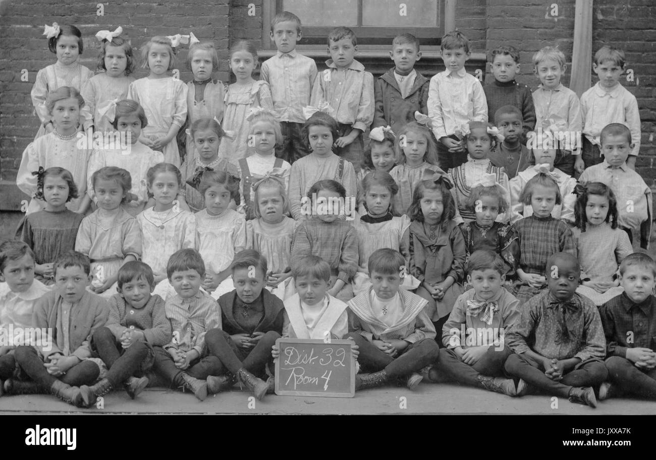 Ganzkörperaufnahme von Schulkindern, alle sitzen außer der hinteren Reihe, ein afroamerikanischer Junge in der ersten Reihe, mit einem Schild mit dem Namen des Lehrers und Zimmernummer, 1915. Stockfoto