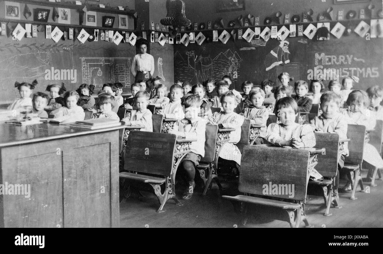 Ganzkörperbild von Schulkindern, die an Schreibtischen sitzen, afroamerikanischer Lehrer im hinteren Teil des Raumes, 'Merry Christmas' auf der Tafel, 1920. Stockfoto