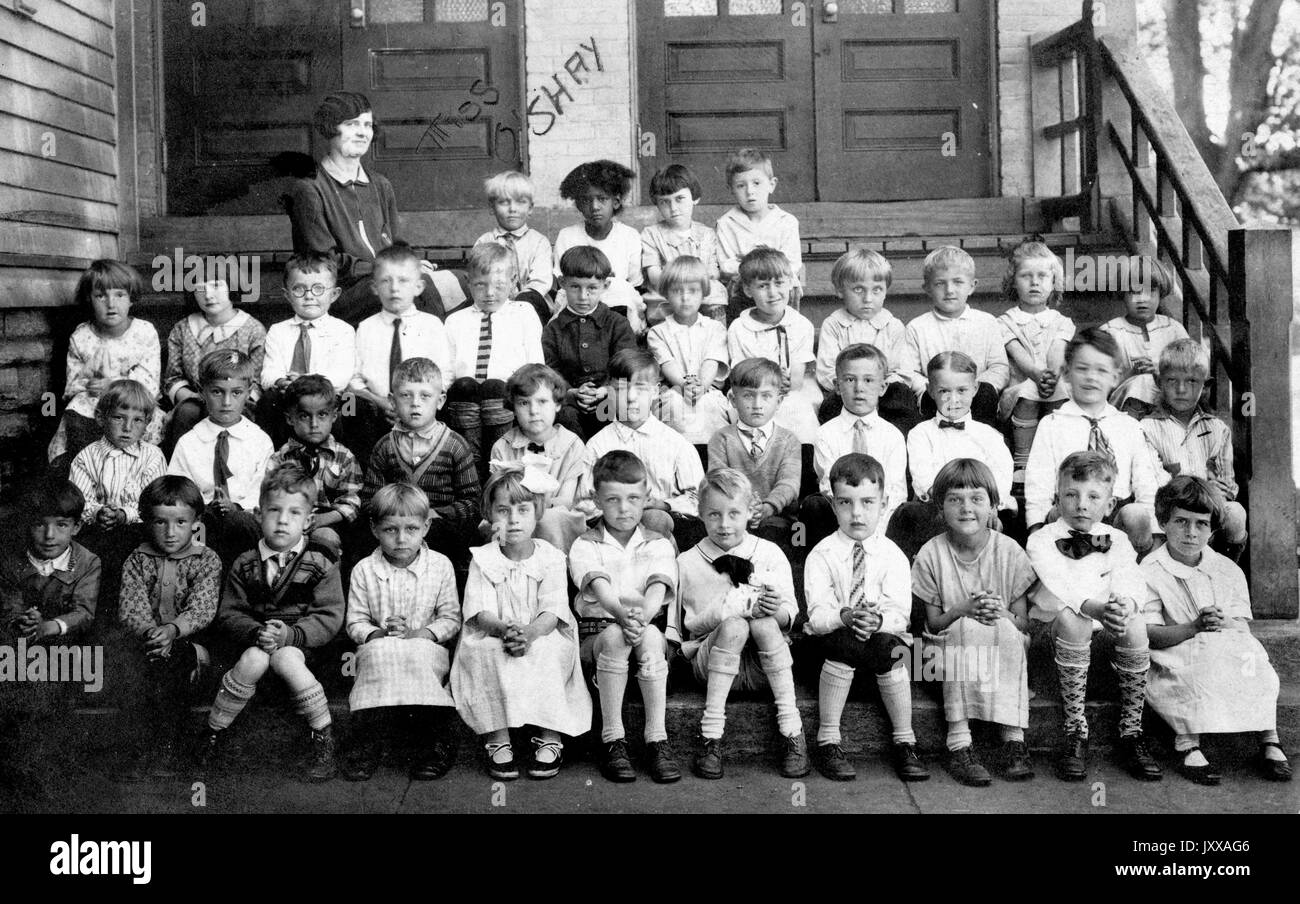 Klassenporträt von 38 jungen Schülern mit Lehrerin, alle weiß bis auf ein afroamerikanisches Mädchen, in Anzügen und Kleidern, auf Treppen vor der Schule sitzend, neutrale Ausdrucksformen, 1915. Stockfoto