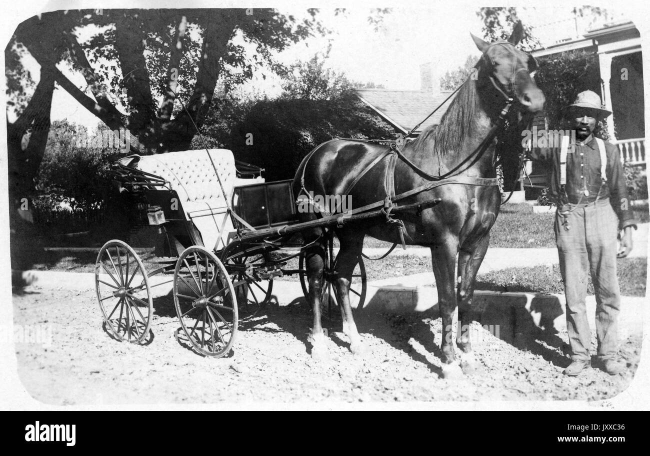 Ganzjähriges Standbild eines reifen afroamerikanischen Mannes mit Pferd und Buggy, in dunklem Hemd, Hosenträger, leichter Hose und Hut, vor Häusern auf Dirt Street stehend, neutraler Ausdruck, 1909. Stockfoto