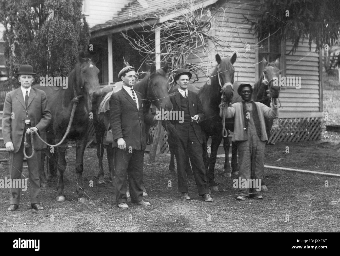 Volle Länge stehen Portraits von drei weißen Männern und einer afrikanischen amerikanischen Mann mit vier Pferden, drei weiße Männer mit dunklen Anzügen, Krawatten, Hüte, afrikanische amerikanische Mann mit dunklen Jacke, Overalls und Hut, stehen vor dem Haus und Bäume, neutrale Ausdrücke, 1920. Stockfoto