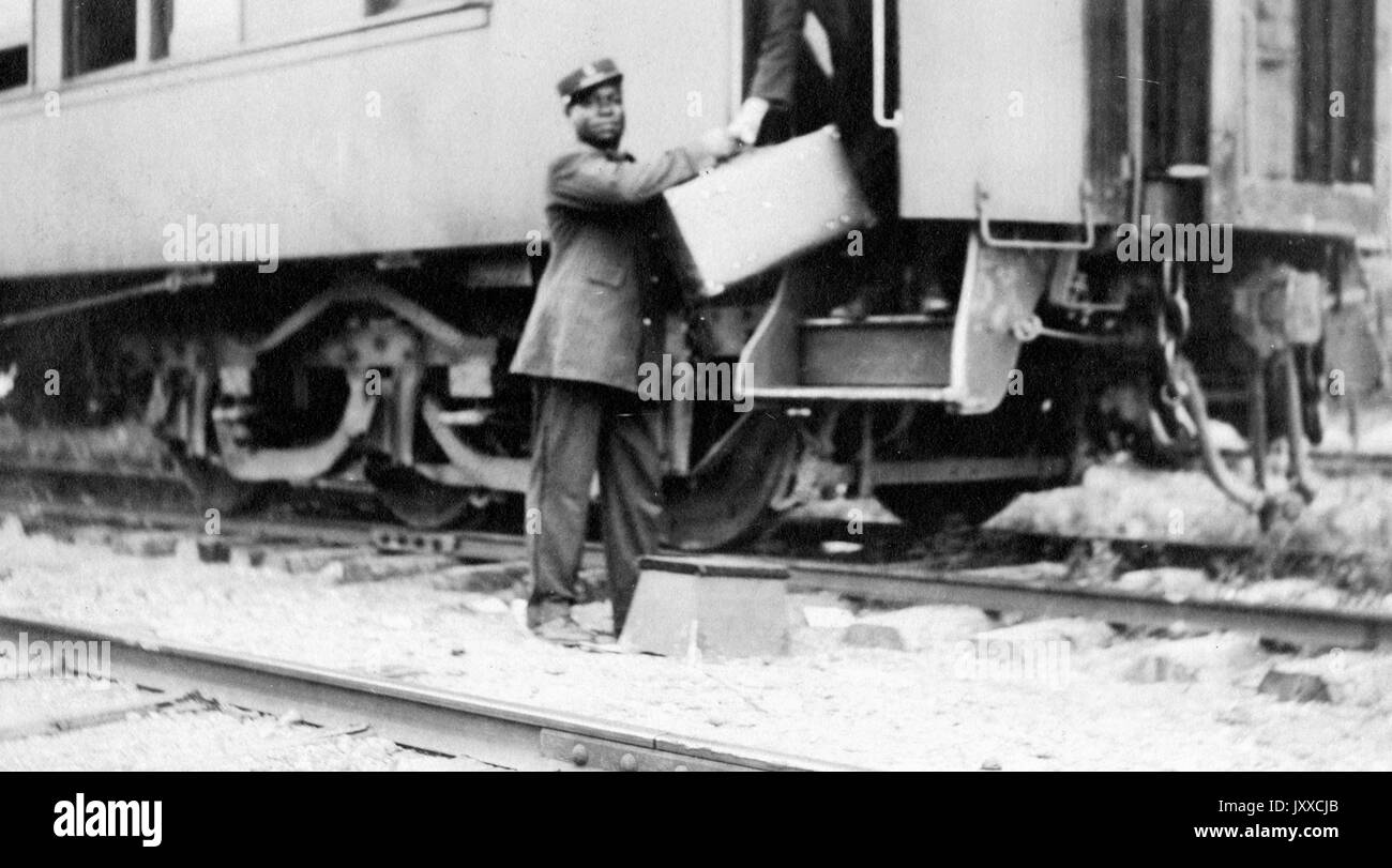 Porträt eines afroamerikanischen Mannes, der in einen Zug einstieg, das Gepäck an Bord übergab, einen dunklen Anzug und einen dunklen Hut trug, zwischen den Gleisen stand, 1920. Stockfoto