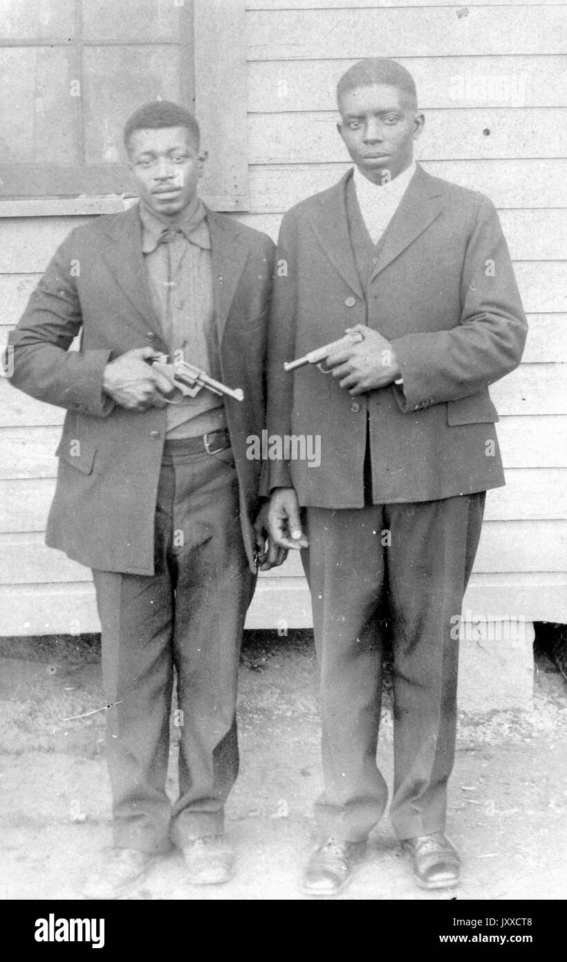 Ganzjähriges Standporträt zweier afroamerikanischer Männer mit neutralen Ausdrücken, die außerhalb eines Gebäudes stehen und mit Handfeuerwaffen aufeinander zeigen, 1915. Stockfoto