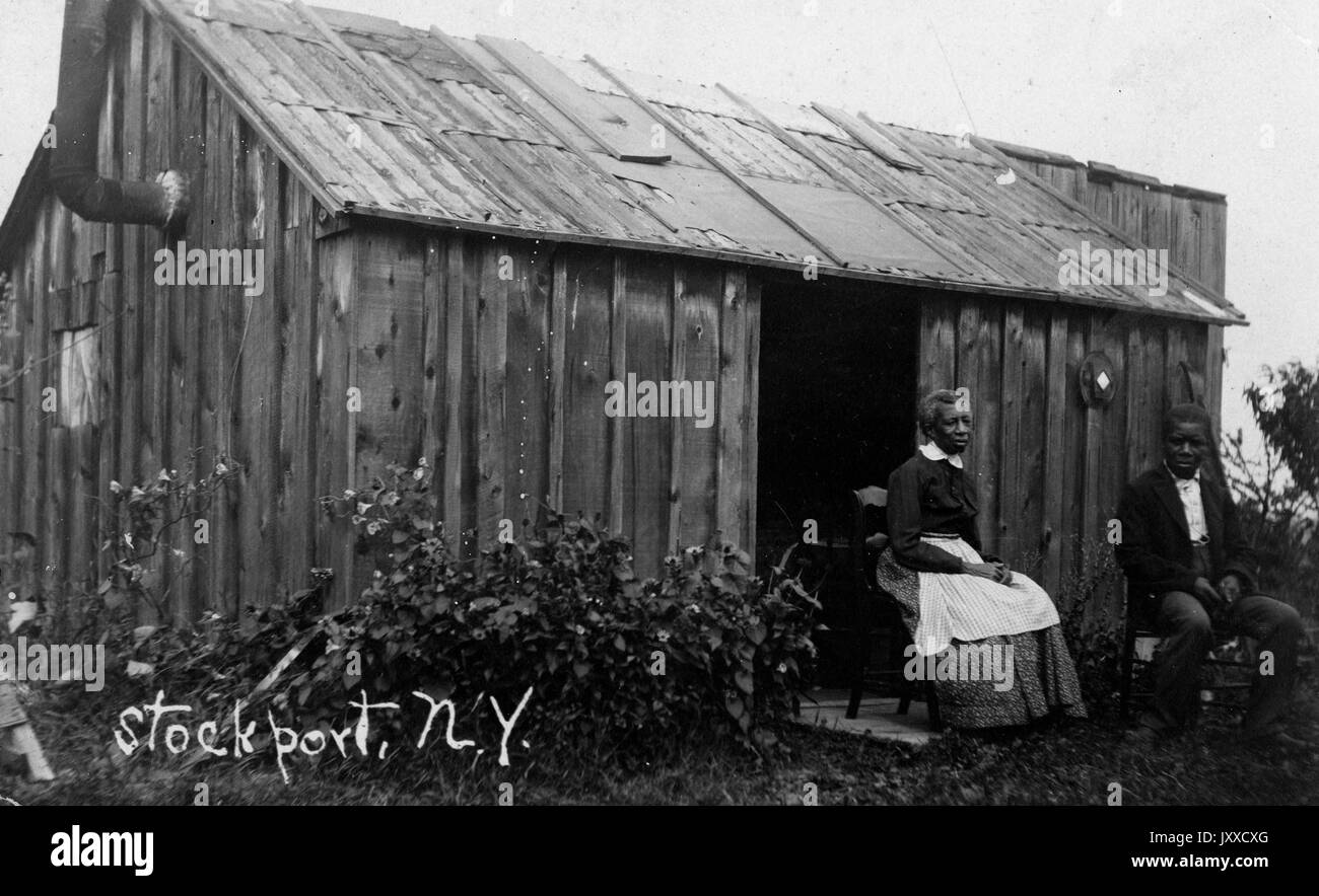 Ein reifer Afroamerikaner und eine reife Afroamerikanerin mit neutralem Ausdruck sitzen außerhalb eines Gebäudes, möglicherweise eines Schuppen, in einer ländlichen Gegend, möglicherweise Stockport, New York, 1909. Stockfoto