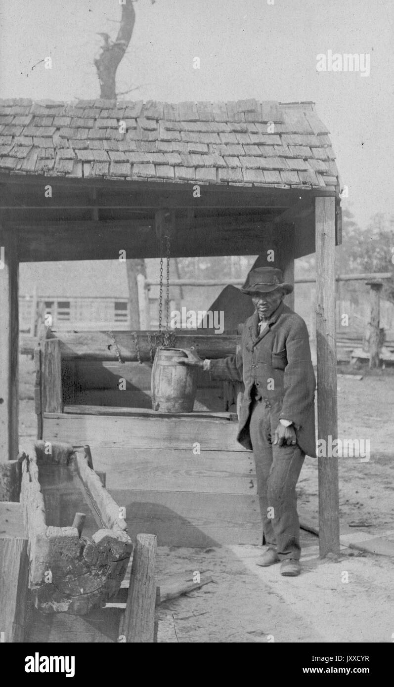 Ein reifer afroamerikanischer Mann mit neutralem Ausdruck sammelt Wasser aus einem Brunnen mit einem Holzfass in einer ländlichen Gegend, 1915. Stockfoto