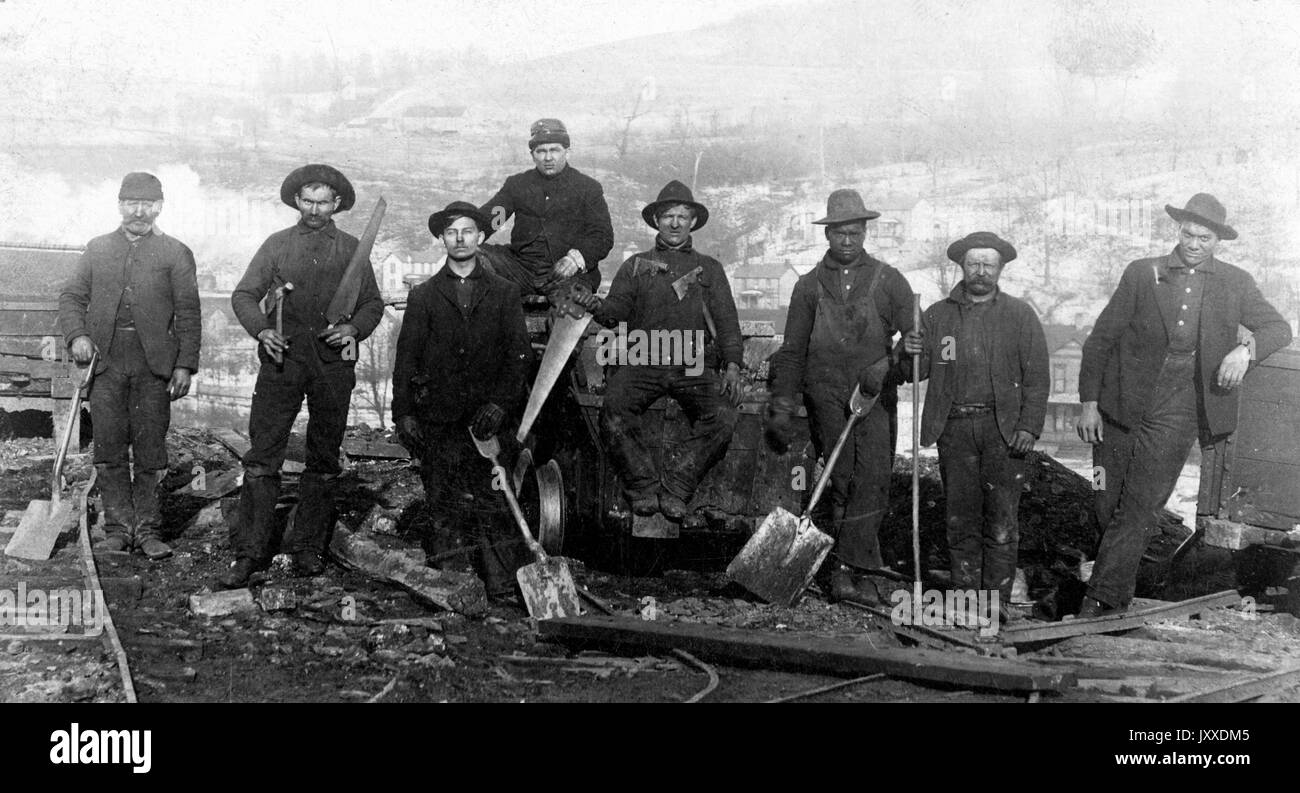 Eine Gruppe amerikanischer Arbeiter mit neutralem Ausdruck steht draußen, mit einer spärlichen Stadt im Hintergrund, inmitten von Schutt auf Zuggleisen, die Werkzeuge wie Schaufeln, Sägen und einen Hammer halten, ein Arbeiter (ganz rechts) lehnt sich an einen Boxwagen, 1925. Stockfoto