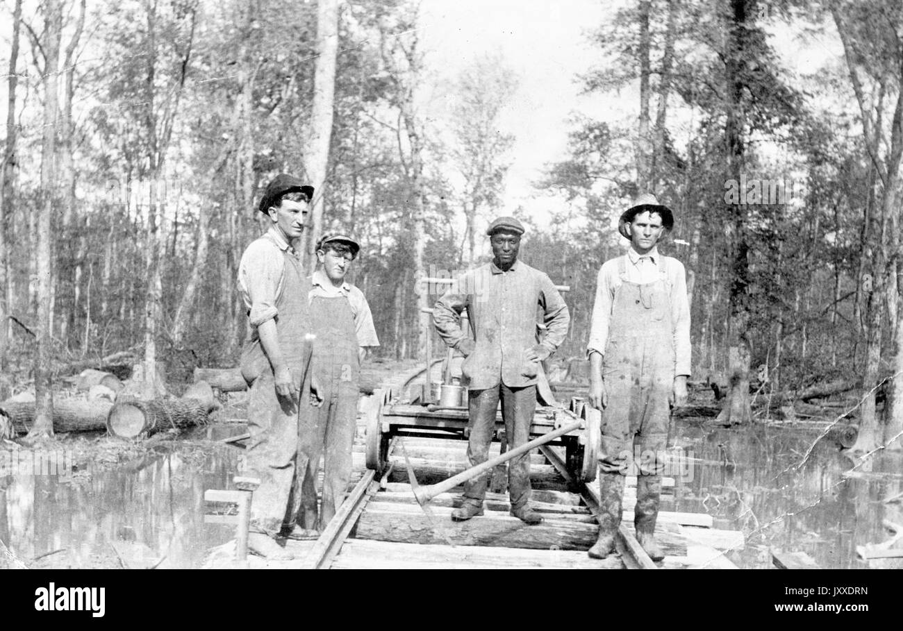 Eine Gruppe amerikanischer Arbeiter mit neutralem Ausdruck steht draußen in einem bewaldeten, sumpfigen Gebiet, auf Schienen, die einen Handwagen tragen, 1920. Stockfoto