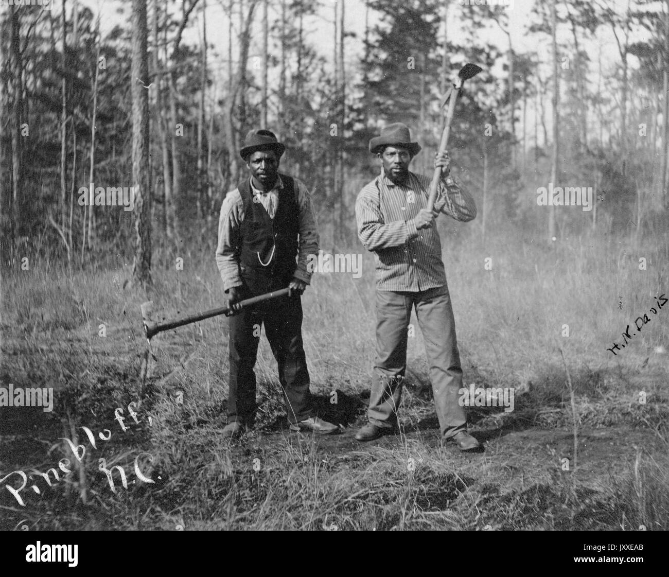 In voller Länge stehend Porträt von zwei afroamerikanischen Arbeiter mit neutralem Ausdruck, tragen Hüte, Hosen, Schuhe, Button-up-Shirts und eine in einem Anzug Weste, stehen in einem gerodeten Waldgebiet mit Äxten, gestellt, als ob sie bei der Arbeit, 1920. Stockfoto
