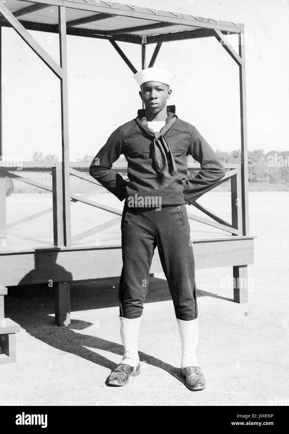 Portrait einer afrikanischen amerikanischen US Navy Sailor stand vor einer hölzernen Plattform, er trägt einen dunklen Sailor uniform und einem hellen Sailor hat, die Arme sind gebeugt und seine Hände sind hinter seinem Rücken, 1920. Stockfoto
