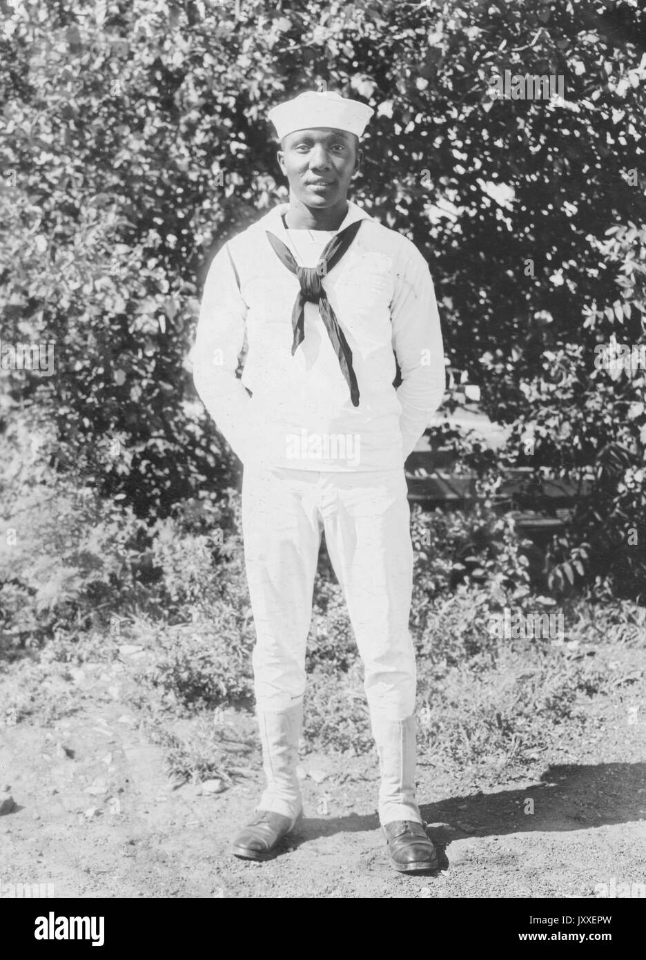 Porträt eines African American US Navy Sailor vor Gebüsch stehend, Hände sind hinter seinem Rücken versteckt und er trägt eine helle Sailor Uniform, 1920. Stockfoto