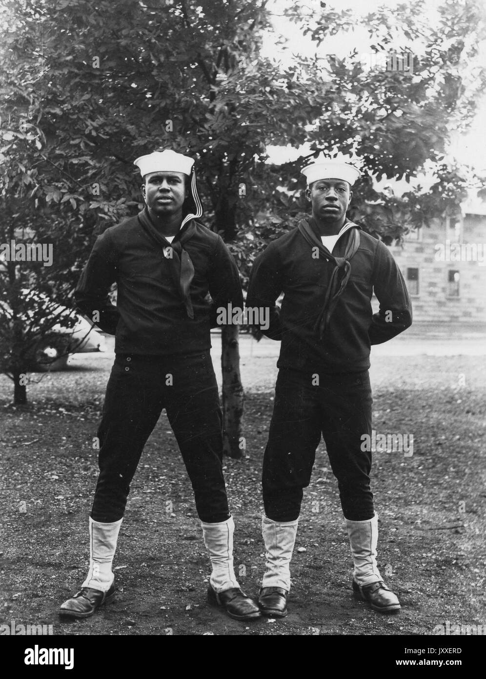 Porträt zweier afroamerikanischer US Navy-Matrosen, die vor einem Baum stehen, beide tragen dunkelfarbige Sailor-Uniformen und helle Sailor-Hüte, beide haben gebeugte Arme und hinter dem Rücken, 1920. Stockfoto