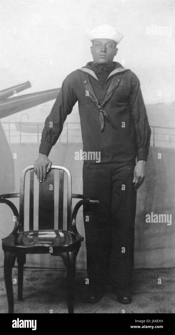 Porträt eines afroamerikanischen US Navy Seemann vor einem Wandbild eines Schiffes mit Kanonen, er trägt eine dunkelfarbige Sailor Uniform und einen hellen Sailor Hut, eine Hand ist an seiner Seite und die andere lehnt sich auf die Rückseite eines Stuhls, 1920. Stockfoto