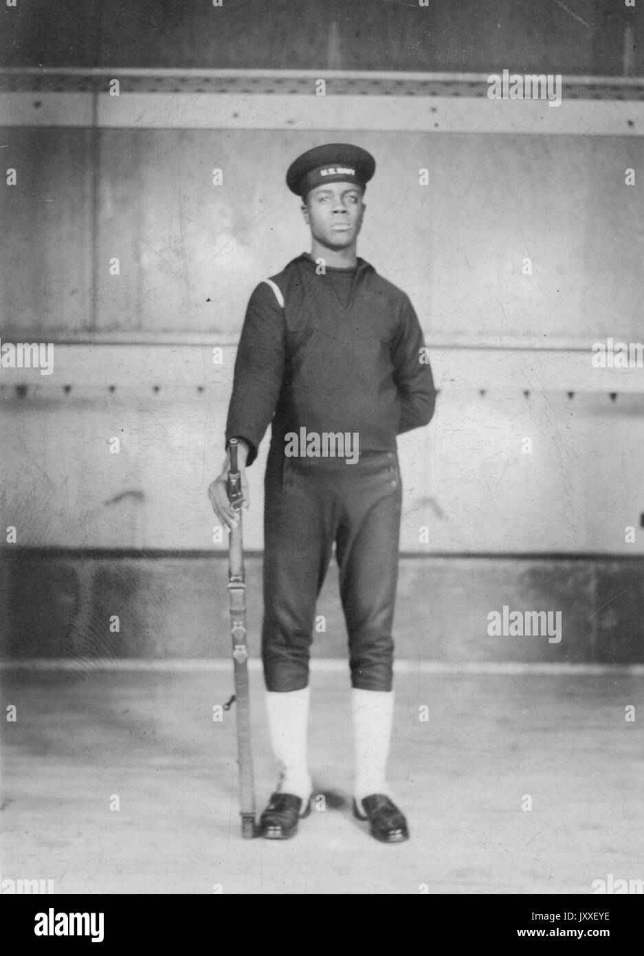 Portrait einer afrikanischen amerikanischen US Navy Sailor stand vor einem großen Schiff, er trägt einen dunklen Sailor Uniform, einen Arm hinter den Rücken und die andere ist im Besitz einer Feuerwaffe durch seine Seite, 1920. Stockfoto
