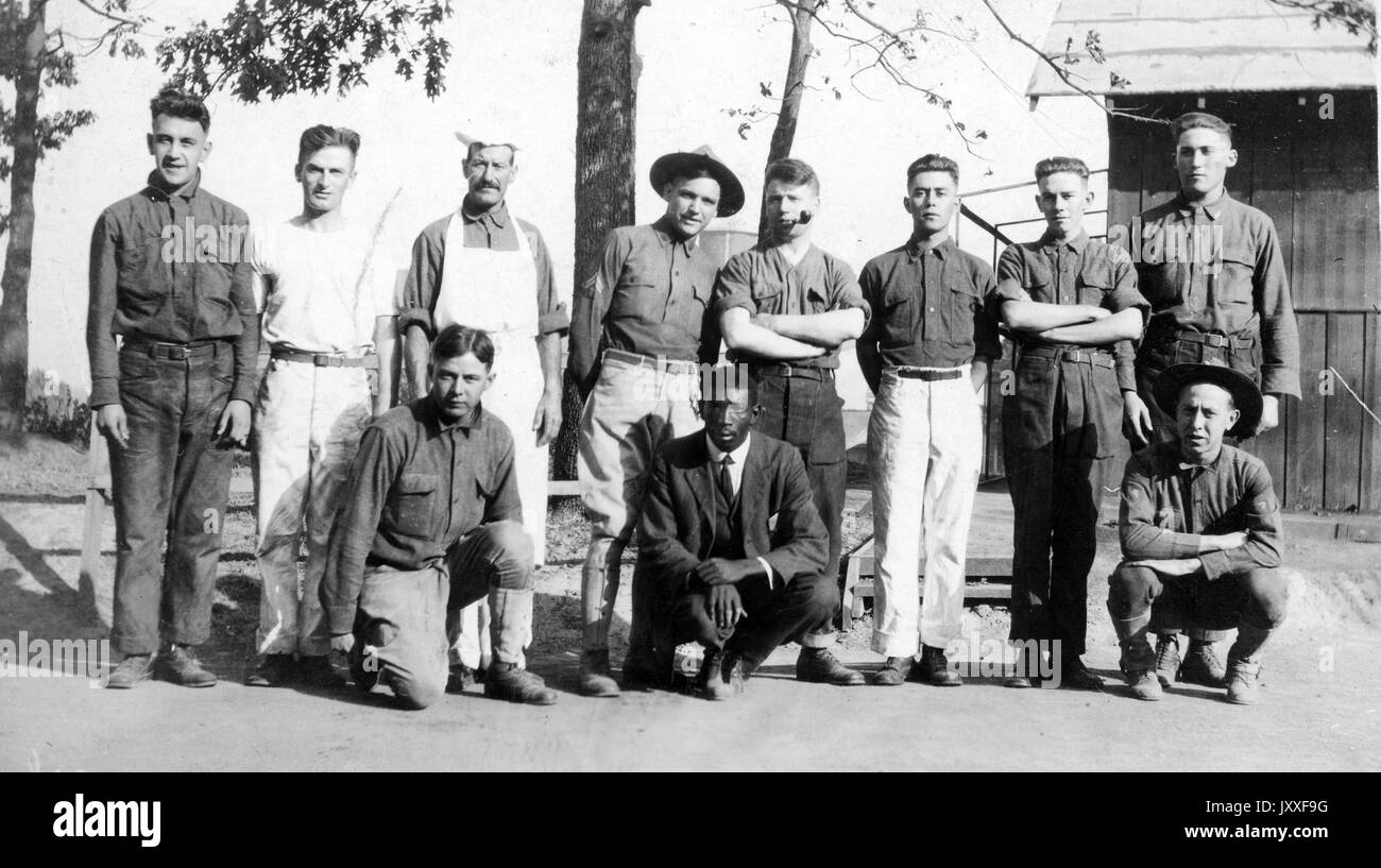 Ein afroamerikanischer Mann hocket mit zwei weißen Männern, hinter ihnen steht eine Linie weißer Männer, der afroamerikanische Mann ist in einem Anzug gekleidet und alle anderen Männer sind jeweils anders gekleidet, sie stehen vor einem Zaun und einer Scheune, 1917. Stockfoto