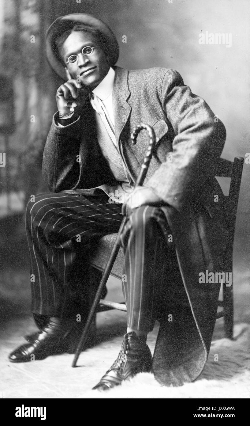 Sitzendes Portrait eines reifen afroamerikanischen Mannes, mit dunklem Mantel, dunkelgestreifter Hose, hellem Hemd, Krawatte, Hut und Klassen, Gehstock haltend, neutraler Ausdruck, 1920. Stockfoto