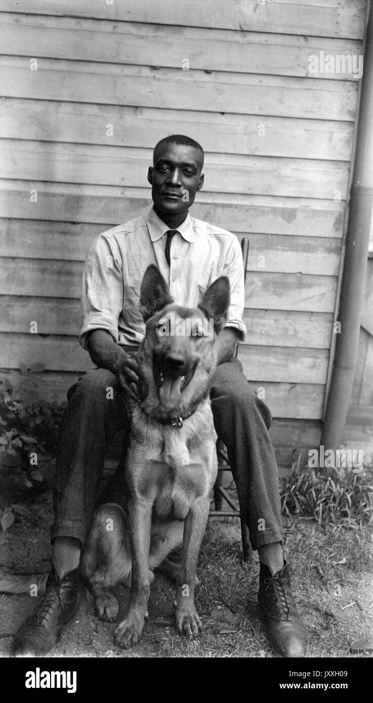 Ganzkörperportrait des reifen afroamerikanischen Mannes, trägt helles Hemd und dunkle Hose, sitzt draußen auf Stuhl vor dem Haus mit Schäferhund sitzen, neutraler Ausdruck, 1925. Stockfoto