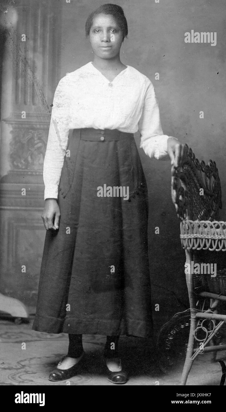 Porträt einer afroamerikanischen Frau, die vor einem Wandbild steht, die linke Hand auf der Rückseite eines Korbsessels, der rechte Arm neben ihr, ein langer, dunkler Rock und eine helle Langarmbluse, 1915. Stockfoto