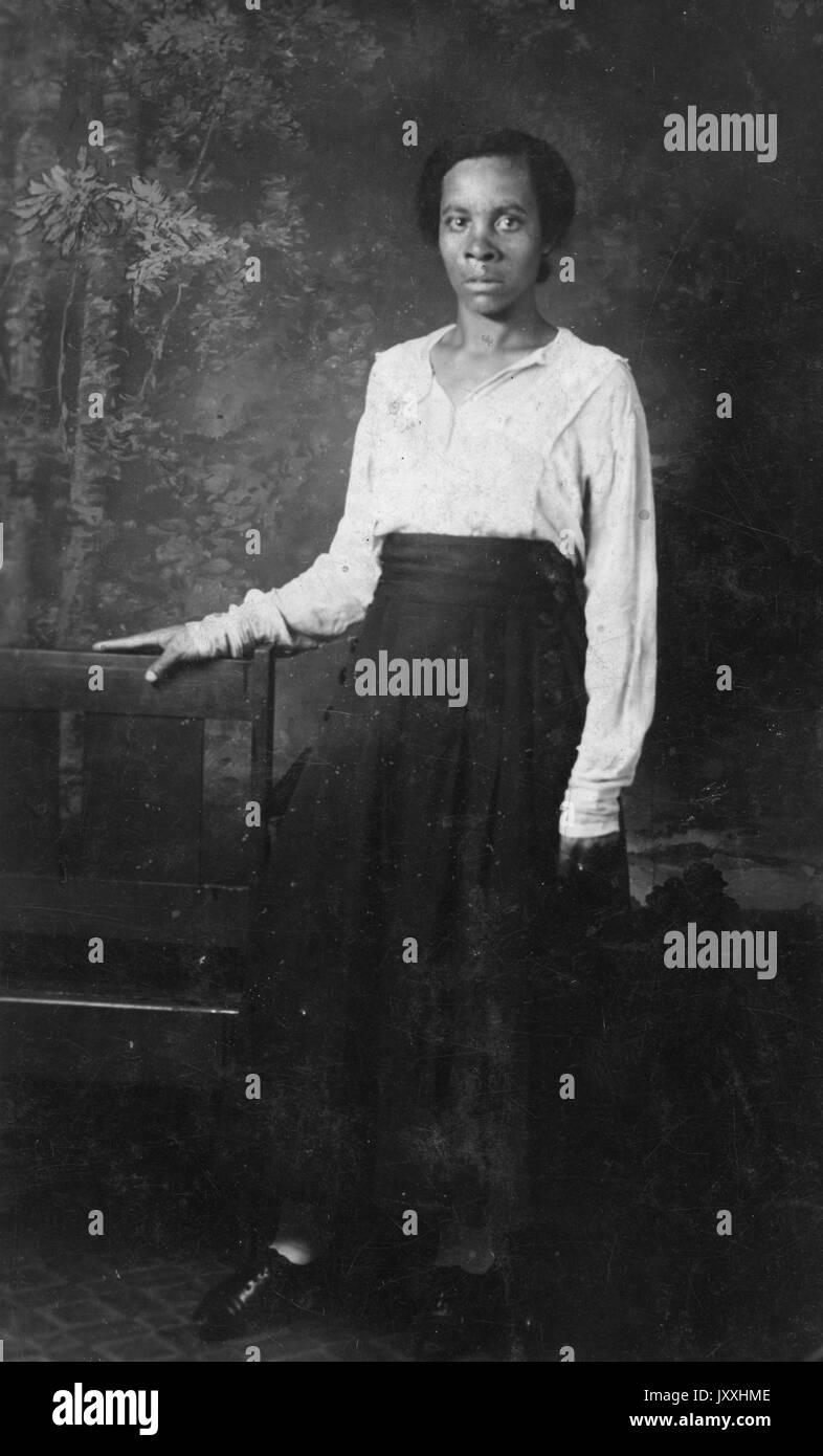 Porträt einer afroamerikanischen Frau, die vor einem Wandbild steht und sich gegen einen Holzstuhl lehnt, ihr rechter Arm ruht ebenfalls auf einem anderen Holzstuhl, ihr linker Arm ist neben ihr, sie trägt einen langen dunklen Rock und eine helle Bluse, 1915. Stockfoto