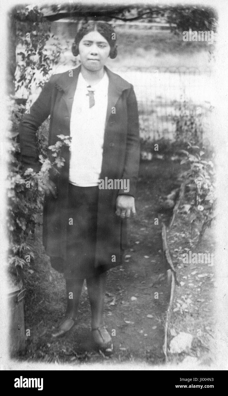 Porträt einer afroamerikanischen Frau, die vor einem Gewässer steht, auf Schmutz steht und links von ihr Büsche trägt, einen dunklen knielangen Rock trägt, eine helle Bluse und einen dunklen langen Mantel darüber, Ihr Haar ist in zwei niedrige Brötchen, 1920, aufgeteilt. Stockfoto