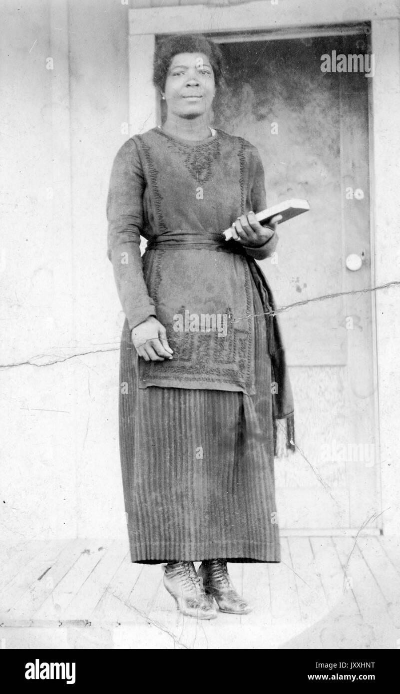 Portrait einer afrikanischen amerikanischen Frau, die vor einer Tür auf einer Veranda, sie trägt eine lange dunkle Kleidung und hält ein Buch in der linken Hand, 1915. Stockfoto