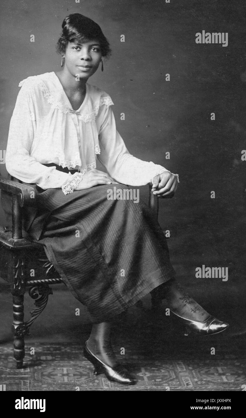 Porträt einer afroamerikanischen Frau, die auf einem geschnitzten Holzstuhl sitzt, die Beine auf den Knien gekreuzt, der linke Arm auf dem Arm des Stuhls ruht und die rechte Hand auf dem Schoß, Sie trägt einen langen dunklen Rock und eine helle Bluse, sie trägt lange Ohrringe, 1915. Stockfoto