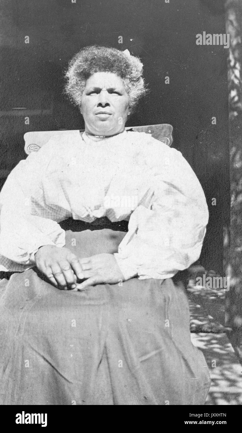 Portrait einer Afrikanische amerikanische Frau in einem Stuhl sitzen mit dem Sonnenlicht auf ihr Gesicht, sie trägt einen langen Rock und eine helle Bluse, sie ist Ihre Augen aufgrund der Sonneneinstrahlung, 1915 schielen. Stockfoto