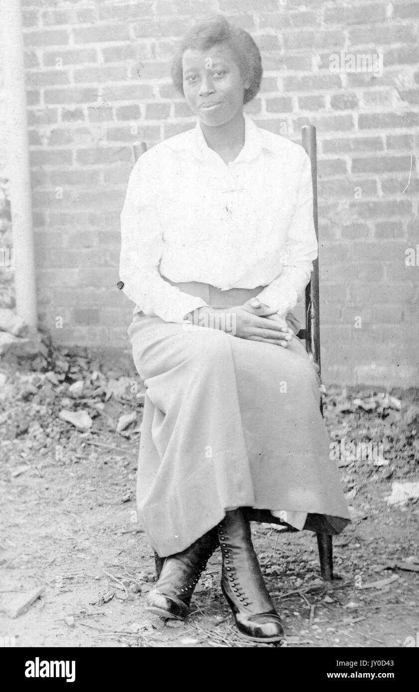 Porträt einer afroamerikanischen Frau, die lächelt und auf einem Stuhl vor einer Ziegelwand sitzt, einen hellen Rock und ein Hemd trägt und ihre Hände auf dem Schoß ruhen, 1915. Stockfoto