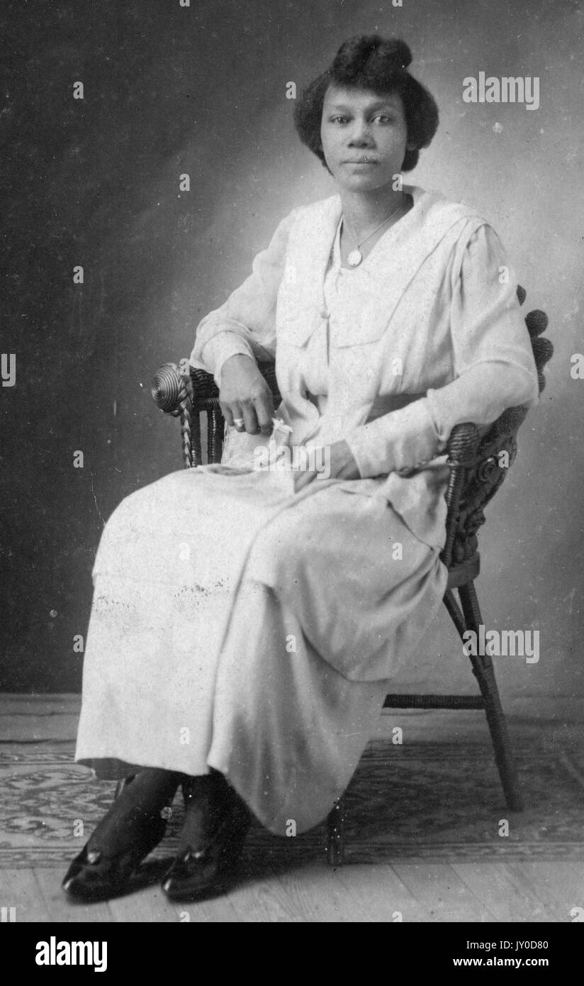 Porträt einer afroamerikanischen Frau, die auf einem Stuhl sitzt, ein langes, helles Kleid und eine passende leichte Jacke trägt, ihre Arme auf den Armen des Stuhls ruhen, ihre Beine sind durch ihre Knöchel gekreuzt, 1915. Stockfoto