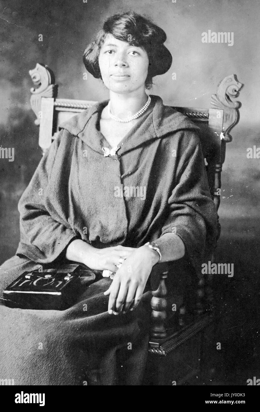 Porträt einer afroamerikanischen Frau, die in einem gebogenen Holzstuhl sitzt, ein langes, dunkelfarbenes Kleid und eine Perlenkette trägt, eine dunkelfarbige Geldbörse ruht auf ihrem Schoß, Ihr rechter Arm ruht ebenfalls auf ihrem Schoß und ihr linker Arm ruht auf dem Arm des Stuhls, 1920. Stockfoto