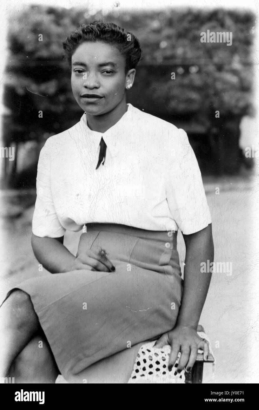 Porträt einer afroamerikanischen Frau, die draußen auf einer Bank mit Bäumen im Hintergrund sitzt, trägt einen knielangen hellen Rock und eine helle Bluse, die Knie werden mit einer Hand im Schoß gekreuzt und die andere auf der Bank ruhend, 1929. Stockfoto