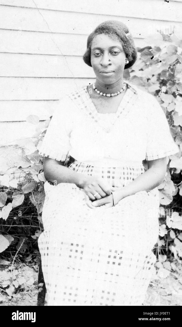 Dreiviertel sitzendes Porträt einer jungen afroamerikanischen Frau mit neutralem Ausdruck, die vor einem Landschaftsbau sitzt und ein leicht gefärbtes gemustertes Kleid, Ohrringe, Halskette und Ring trägt, 1925. Stockfoto