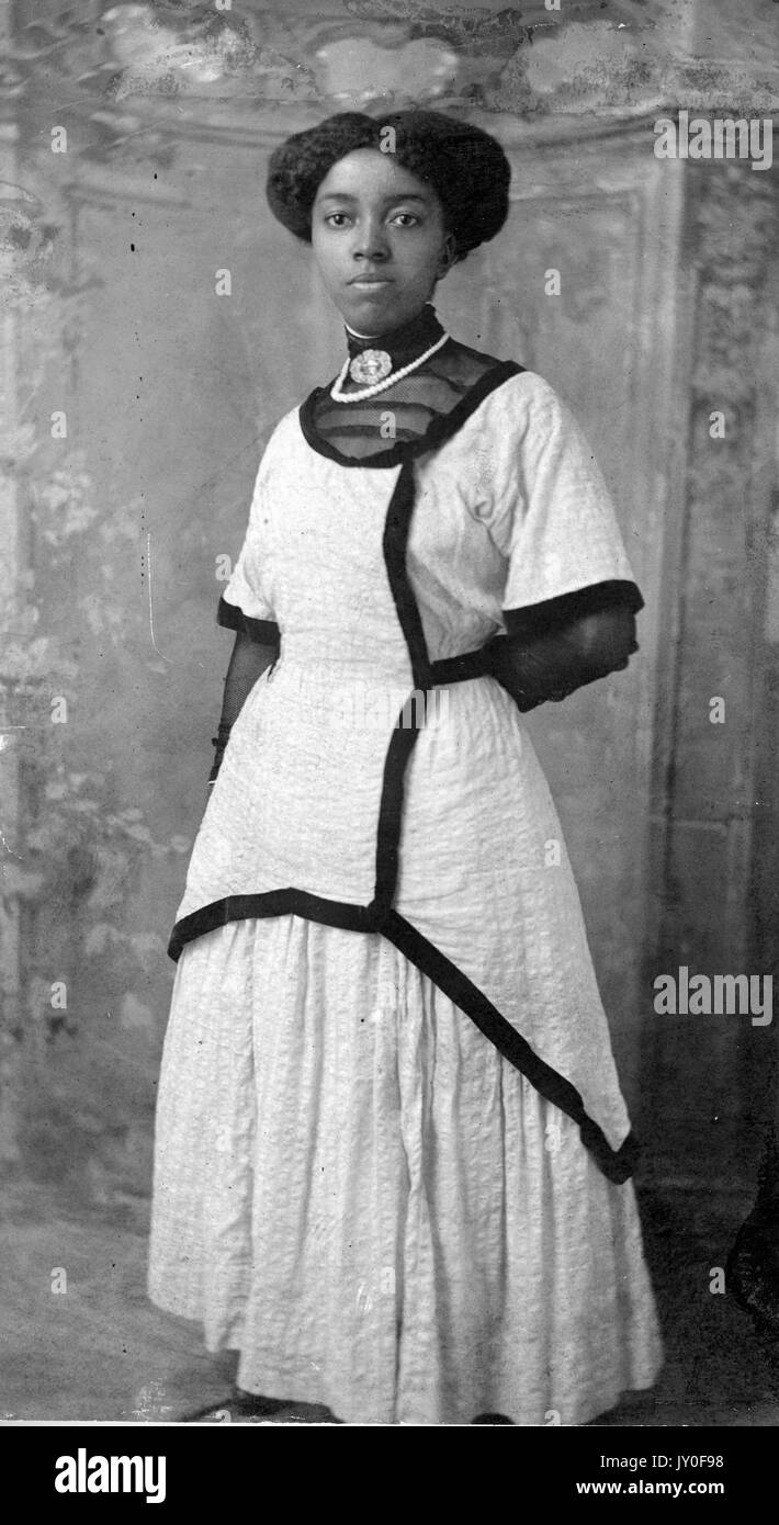 Ganzlanges Standbild einer jungen afroamerikanischen Frau mit neutralem Ausdruck, vor bemaltem Hintergrund, in einem aufwendigen, leicht farbigen Kleid mit dunklem Besatz und Halskette, 1920. Stockfoto